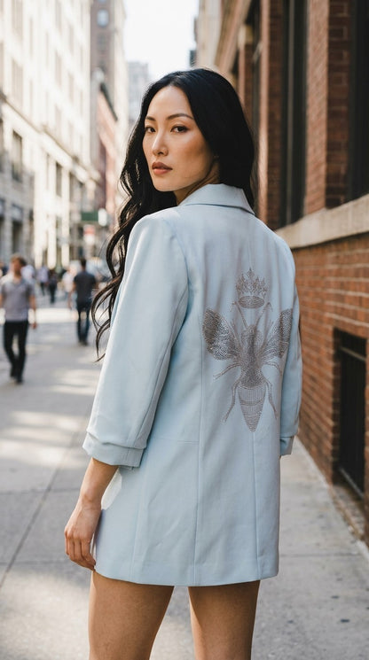 Luxury Clique light blue tailored blazer with a sparkling sequin bee motif on the back, worn by a woman outdoors.