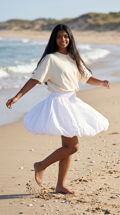 Luxury Clique white high-waisted bubble mini skirt with a smocked waistband worn by a woman on a sandy beach.